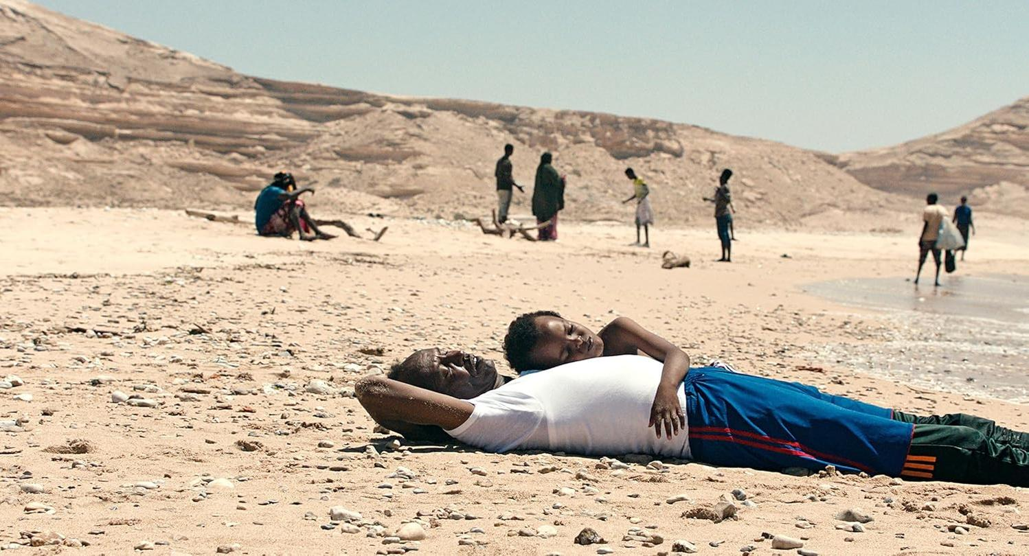 Menschen an einem Sandstrand. Ein Mann und ein Kind liegen. Andere Leute sind am Strand und im Wasser.