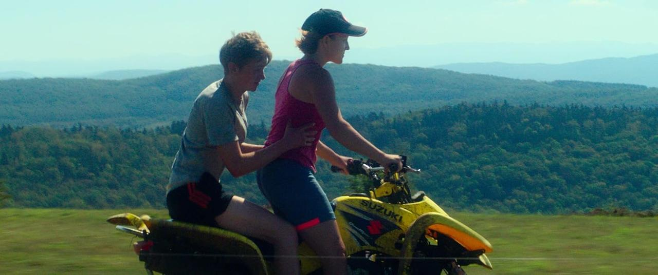 Zwei Personen auf einem gelben ATV auf einer Straße, mit grüner Landschaft im Hintergrund.