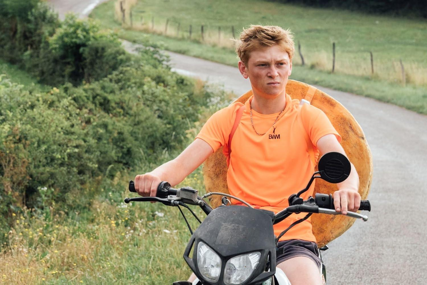 Ein Junge auf einem Motorrad, der einen Schwimmring hält. Er trägt ein orangefarbenes Hemd. Grüner Hintergrund.