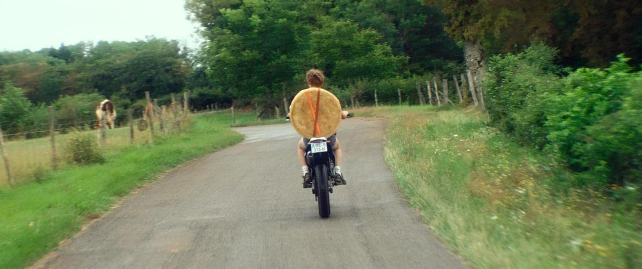 Person auf einem Motorrad mit einem großen runden Gegenstand auf dem Rücken, auf einer Straße.