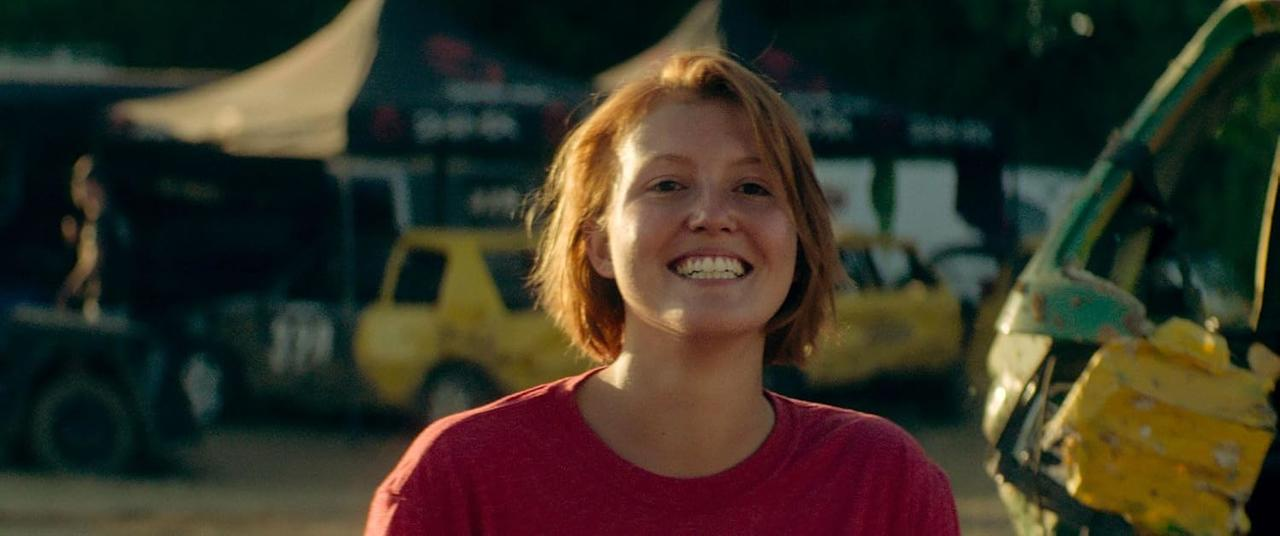 Eine Frau lächelt in die Kamera. Sie hat rotes Haar und trägt ein rotes Hemd. Autos im Hintergrund.