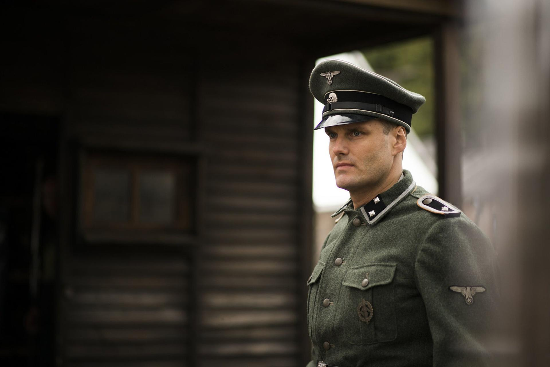 Ein Mann in Nazi-Uniform. Er schaut zur Seite. Er steht vor einem Holzgebäude.