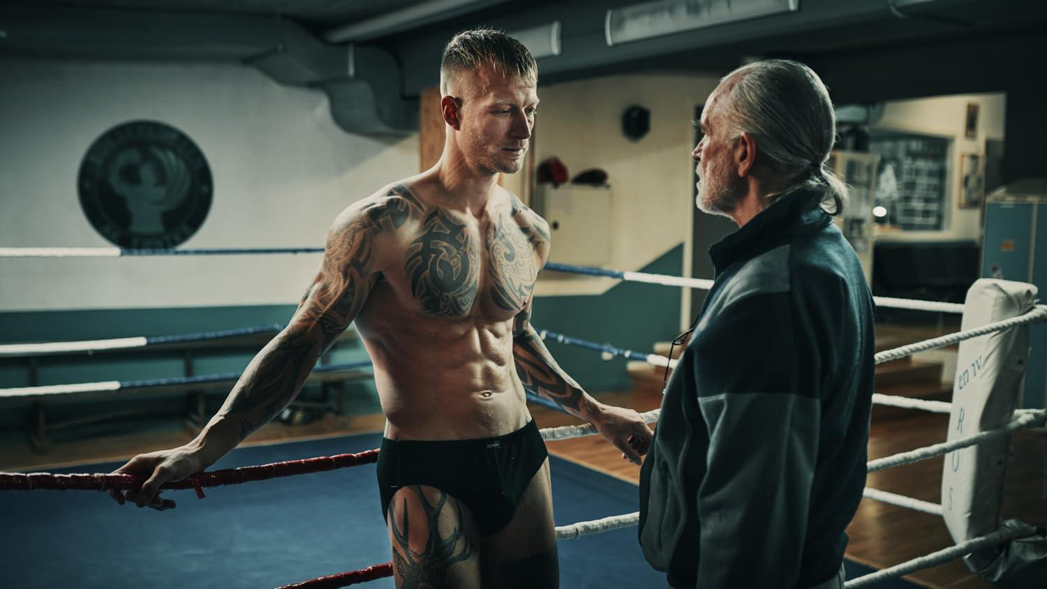 Ein Boxer mit Tätowierungen im Ring, der mit einem Mann in einer Jacke spricht.
