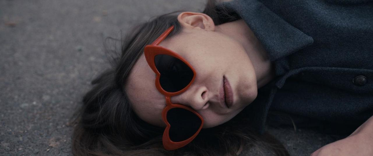 Eine Frau mit herzförmiger Sonnenbrille liegt auf dem Bürgersteig. Ihr Gesicht ist sichtbar und sie schaut in die Kamera.