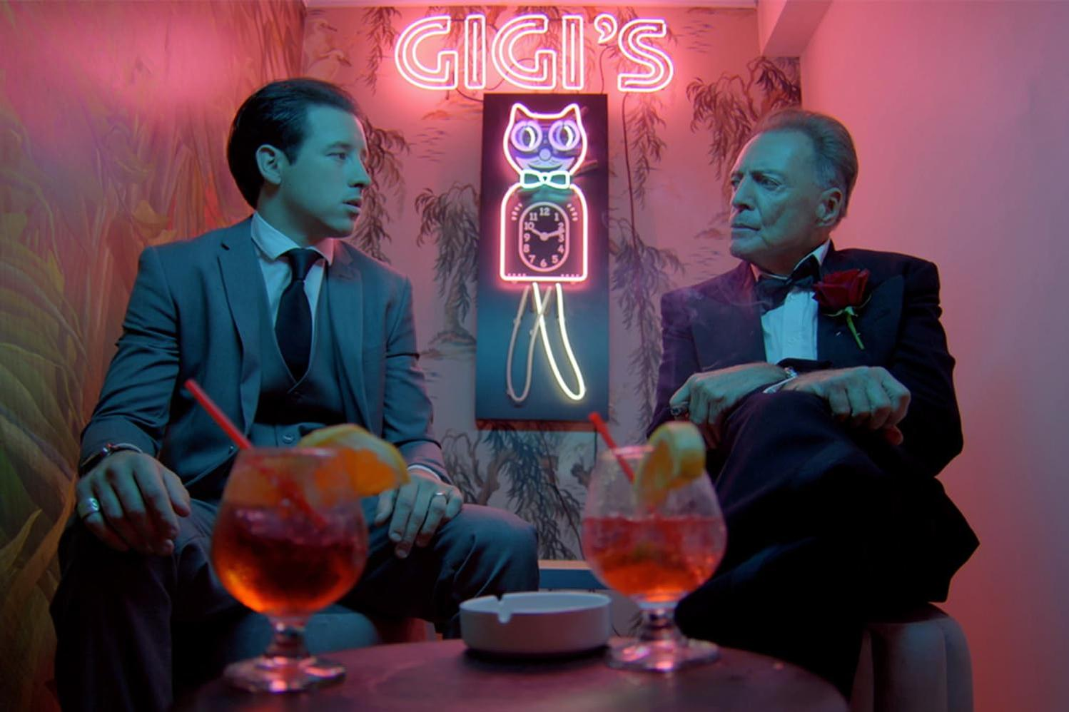 Zwei Männer in einer Bar, Leuchtreklame sagt Gigi's. Cocktails auf dem Tisch.