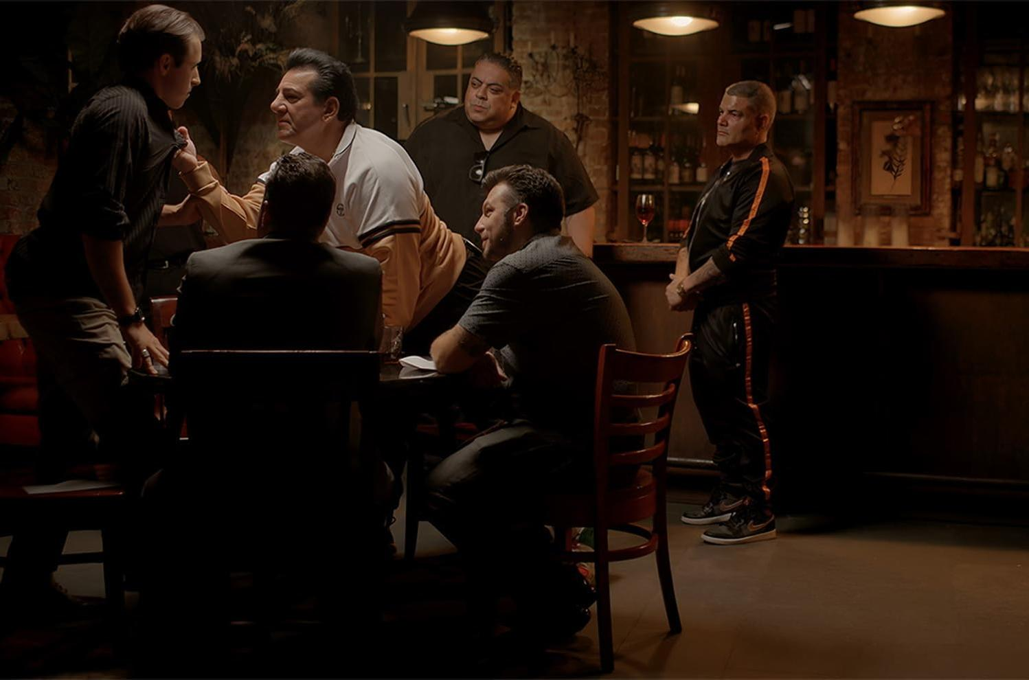 Männergruppe in einer Bar, einige sitzen an einem Tisch, andere stehen. Gedämpfte Beleuchtung, dunkle Atmosphäre.
