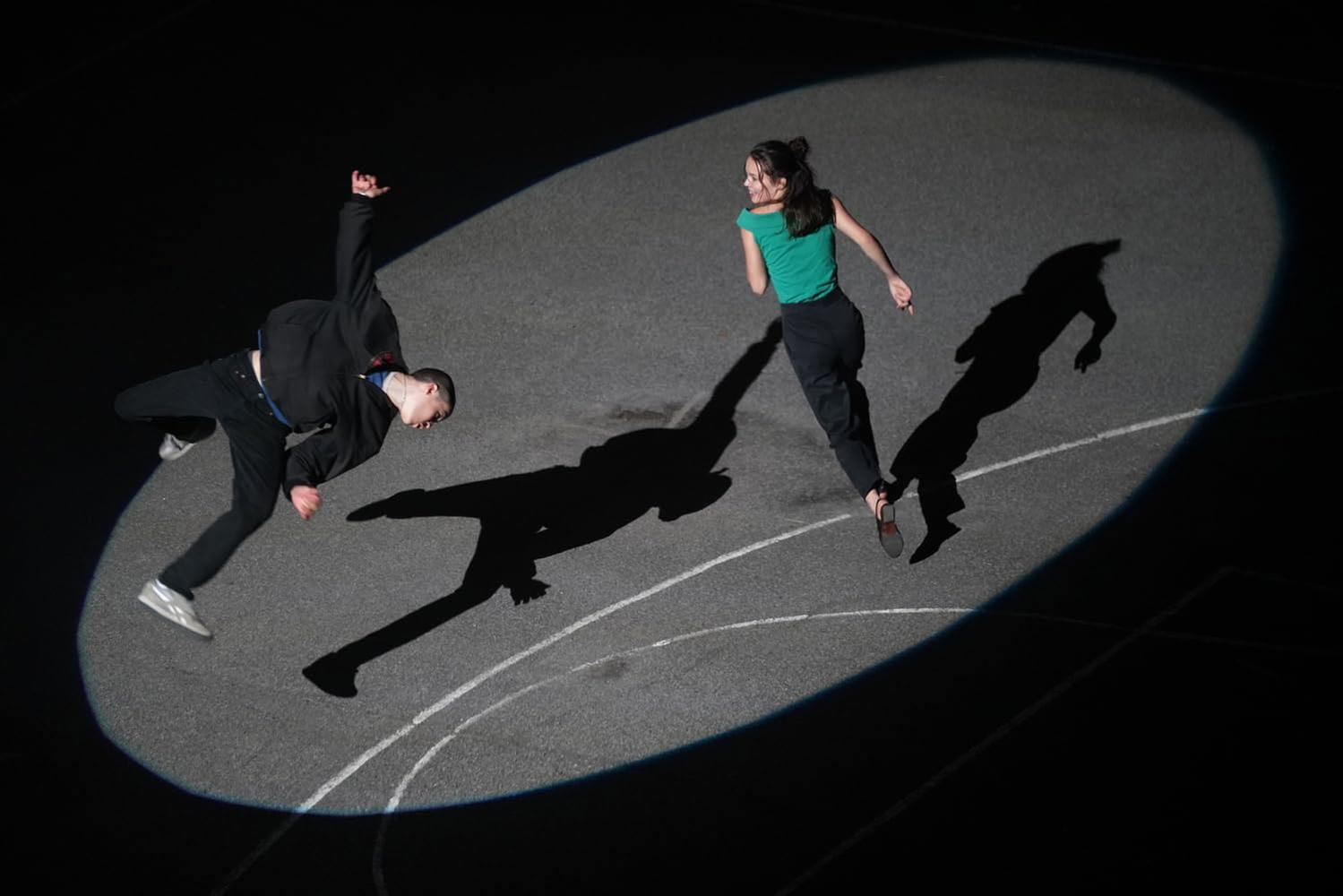 Zwei Tänzer im Scheinwerferlicht. Einer fällt, der andere rennt. Schatten auf dem Boden.