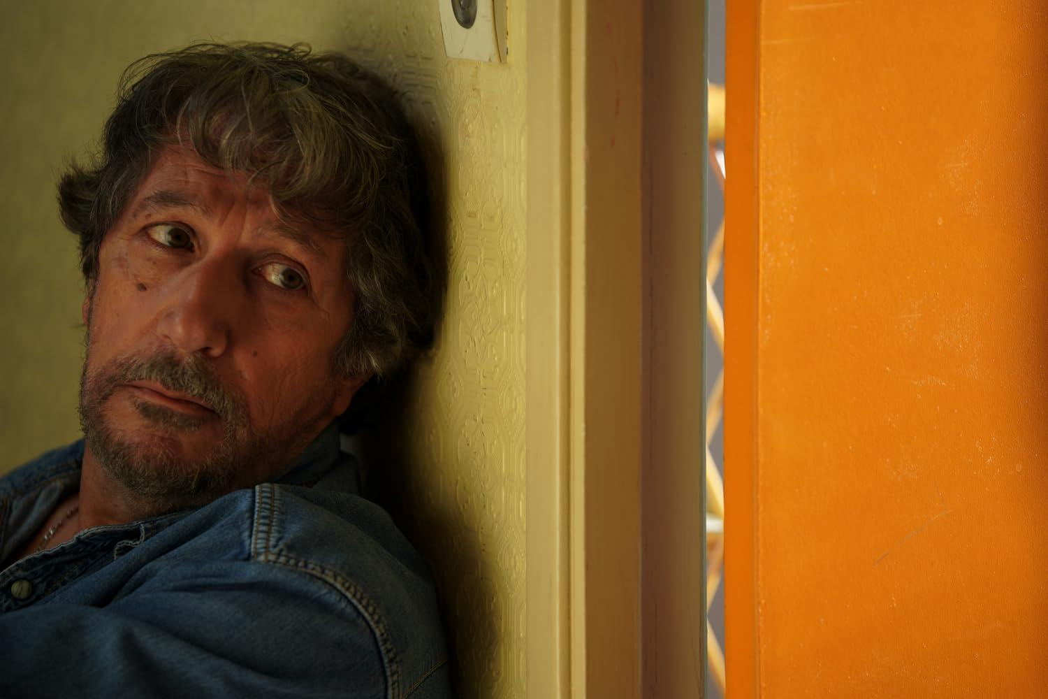 Älterer Mann in Jeanshemd lehnt an gelber Wand und orangefarbenem Türrahmen.