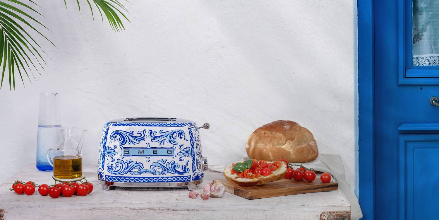 Una escena de cocina con una tostadora SMEG, pan, tomates, aceite de oliva y una puerta azul.