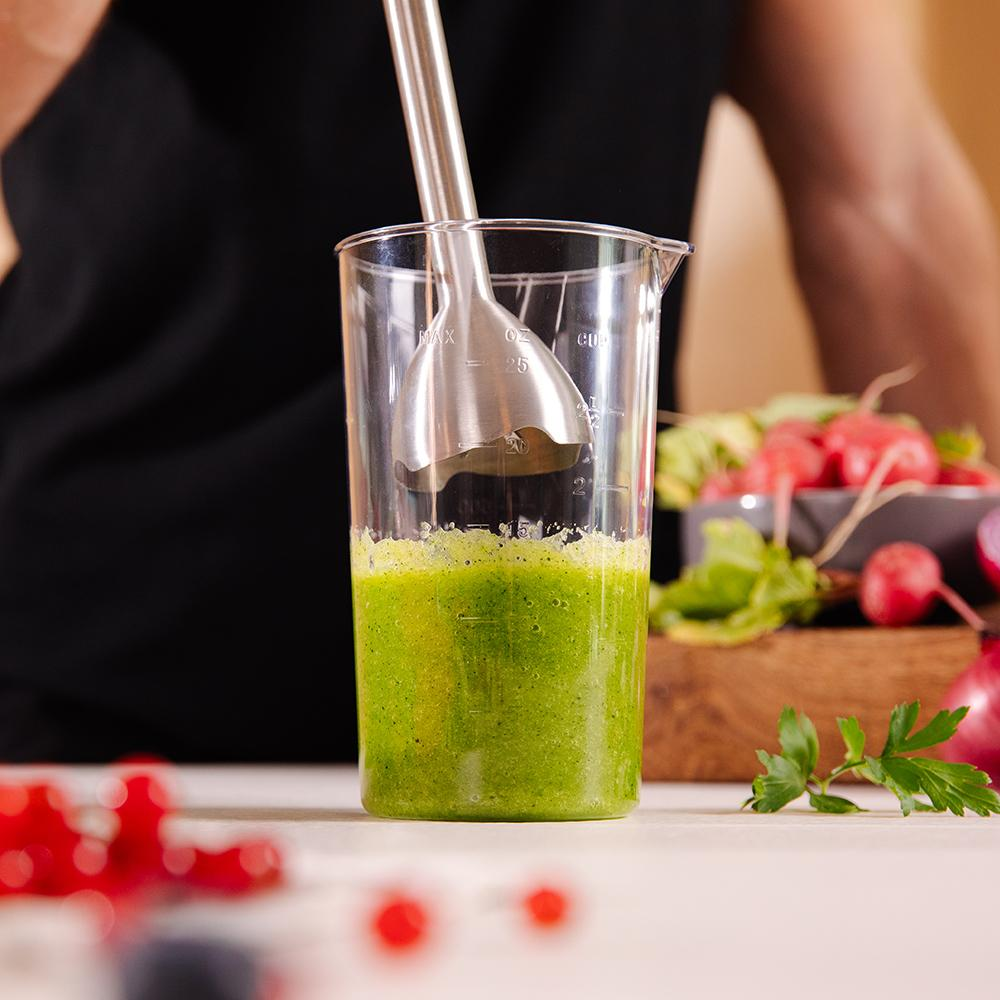 Persona mezclando ingredientes verdes en un vaso transparente, con verduras y batidora.