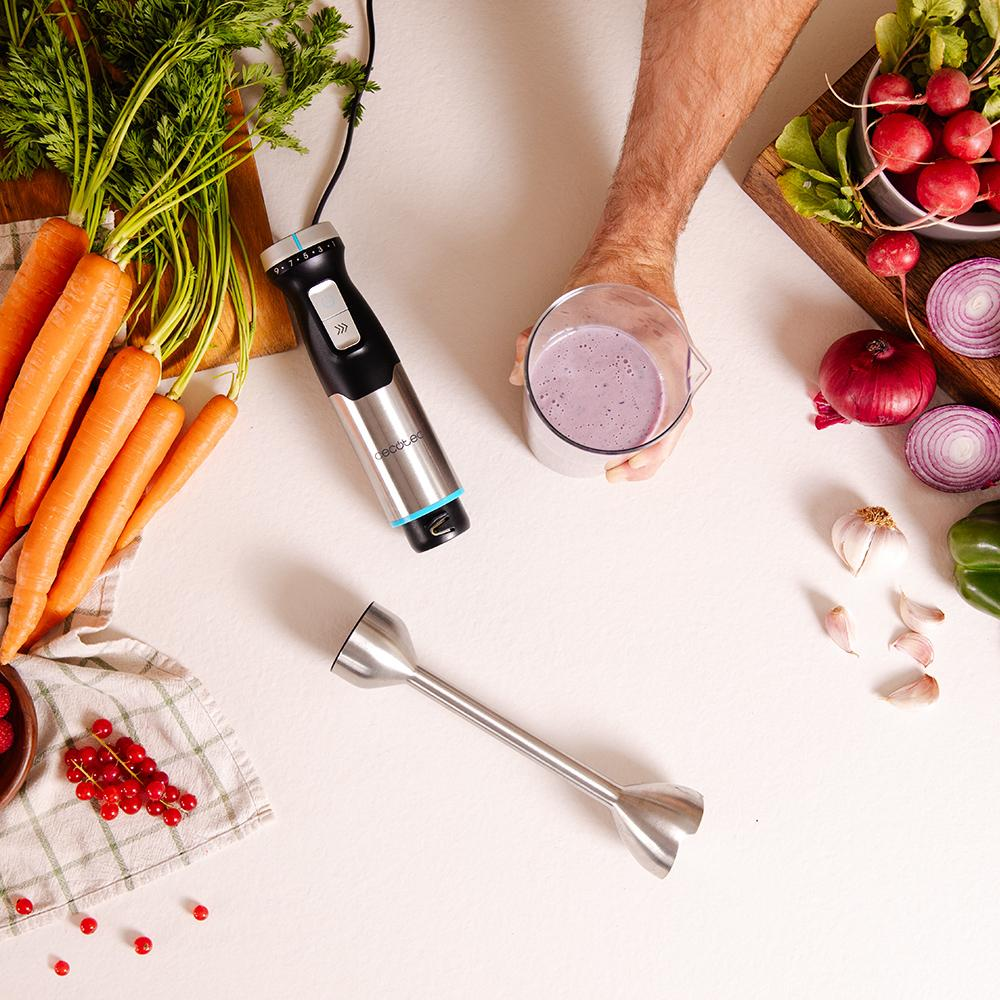 Mano sosteniendo un vaso de batido, batidora y verduras en una mesa.
