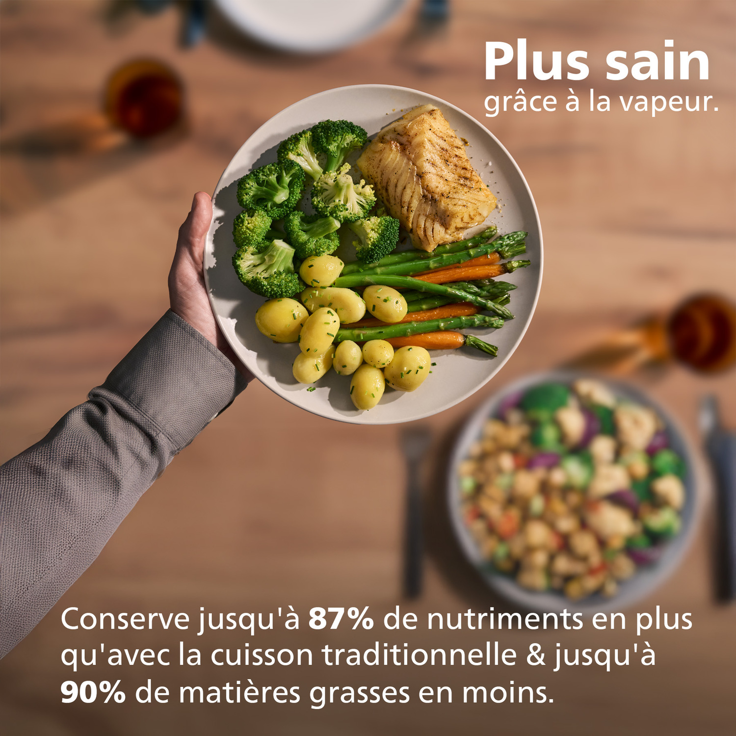 Assiette avec brocolis, asperges, pommes de terre et poisson tenue par une personne. Assiettes floues en fond.