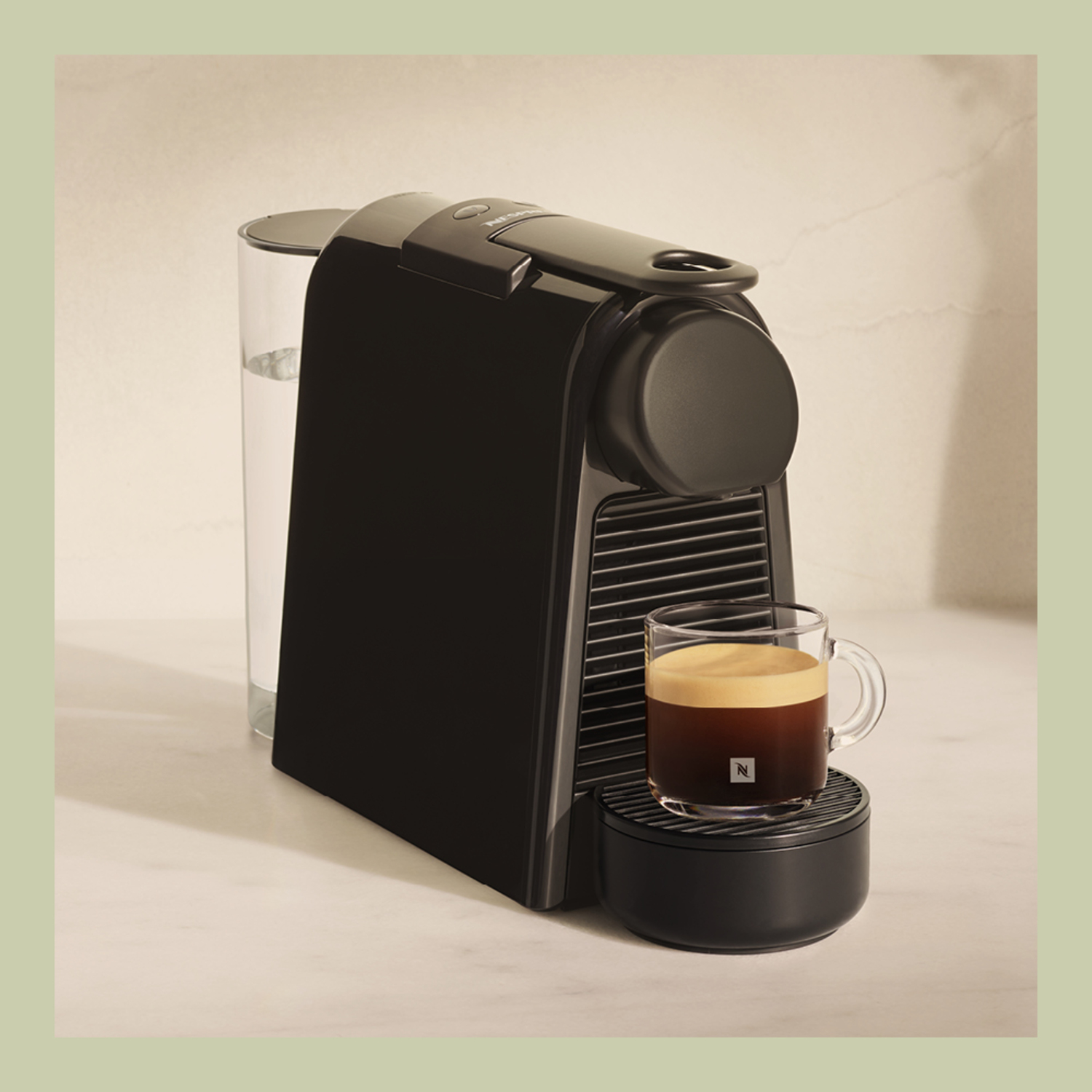 Black Nespresso coffee machine with a glass cup of coffee on a black base, set on a white surface.