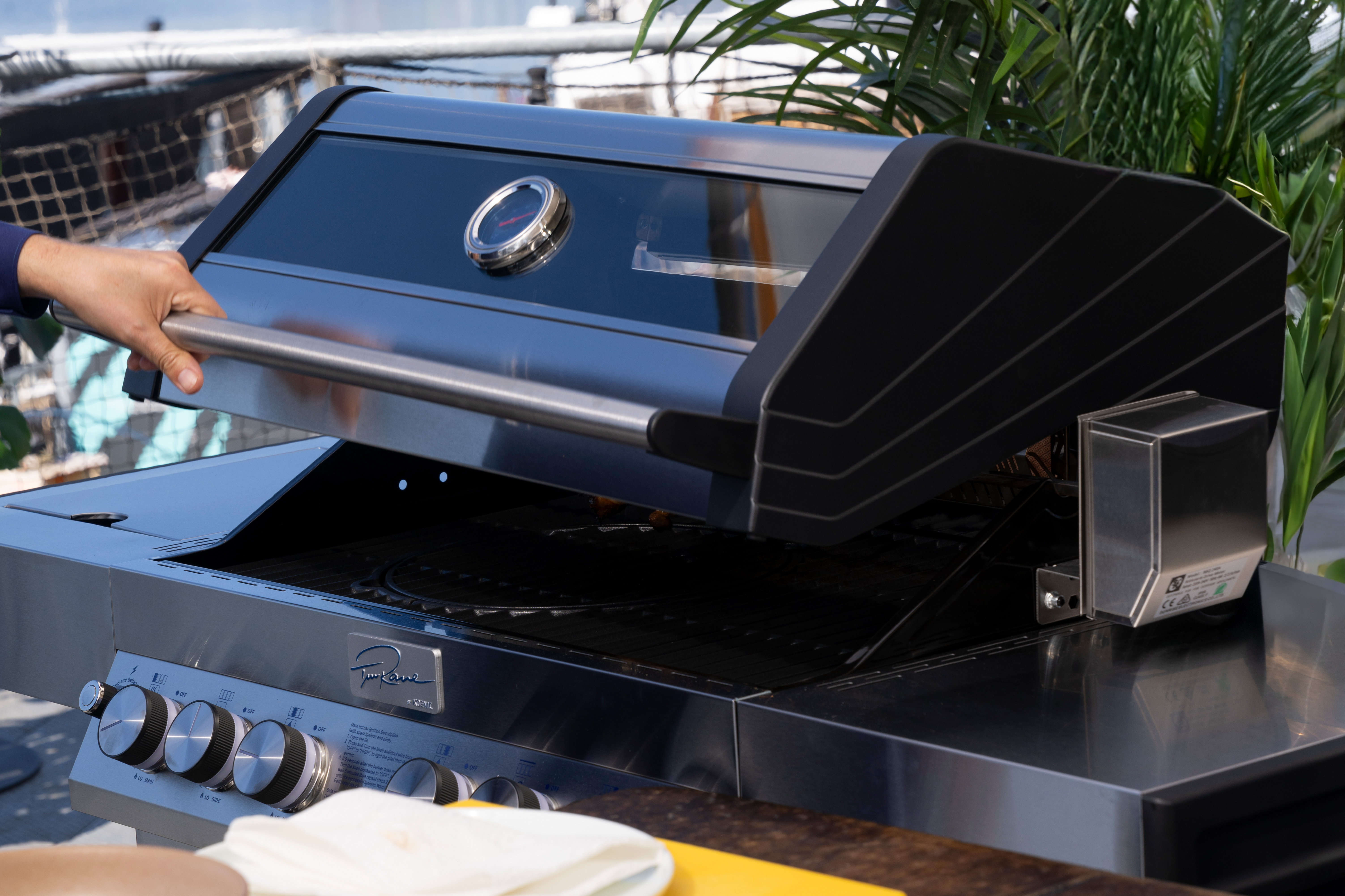Eine Person öffnet einen schwarzen Grill. Der Grill steht auf einer Holzoberfläche in der Nähe von Pflanzen.