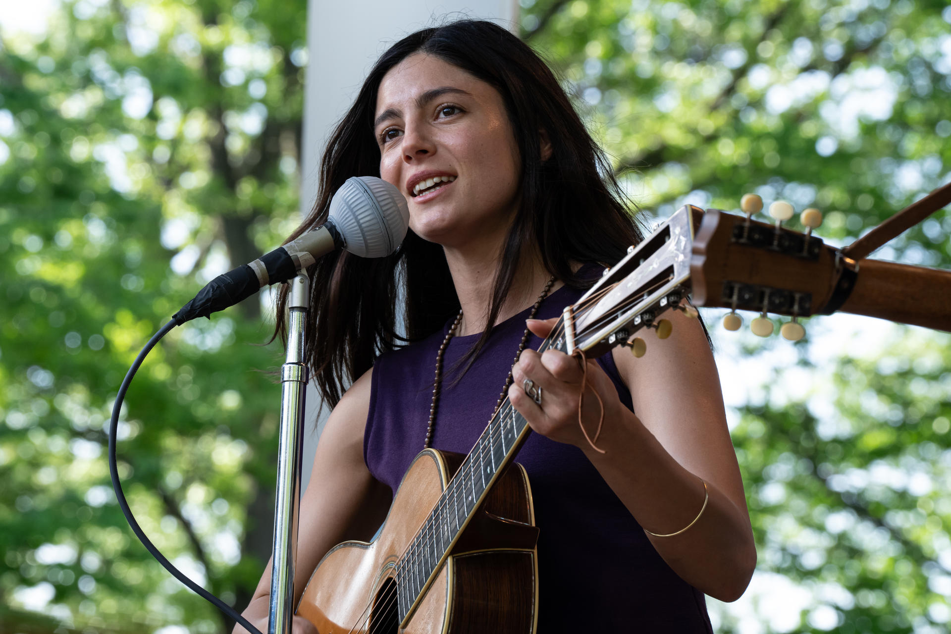 Frau singt und spielt Gitarre im Freien. Sie trägt ein lila Kleid und Halskette, mit Mikrofon.