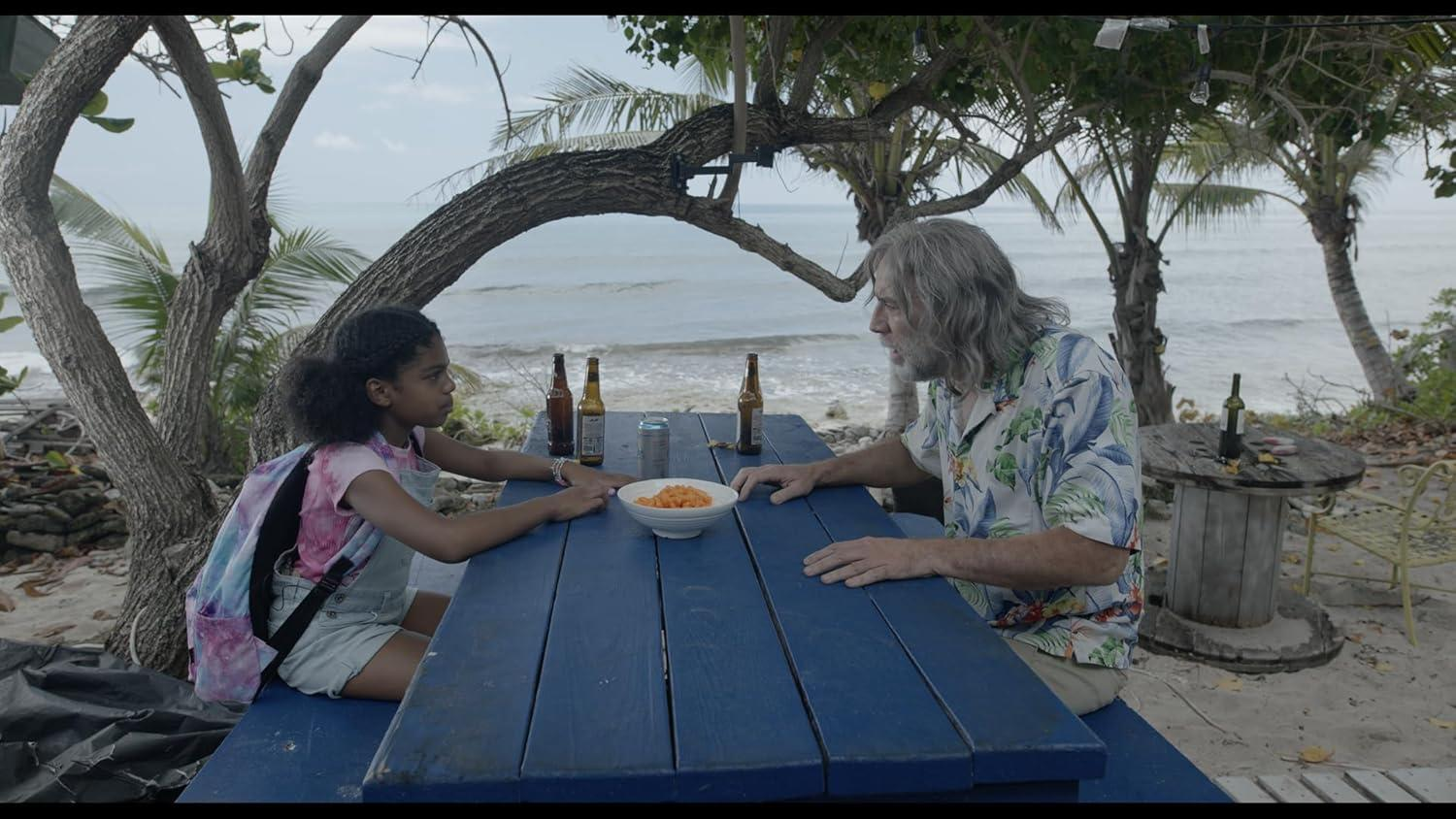 Ein Mädchen und ein älterer Mann sitzen an einem blauen Picknicktisch. Auf dem Tisch stehen Getränke und Snacks. Strand und Bäume im Hintergrund.