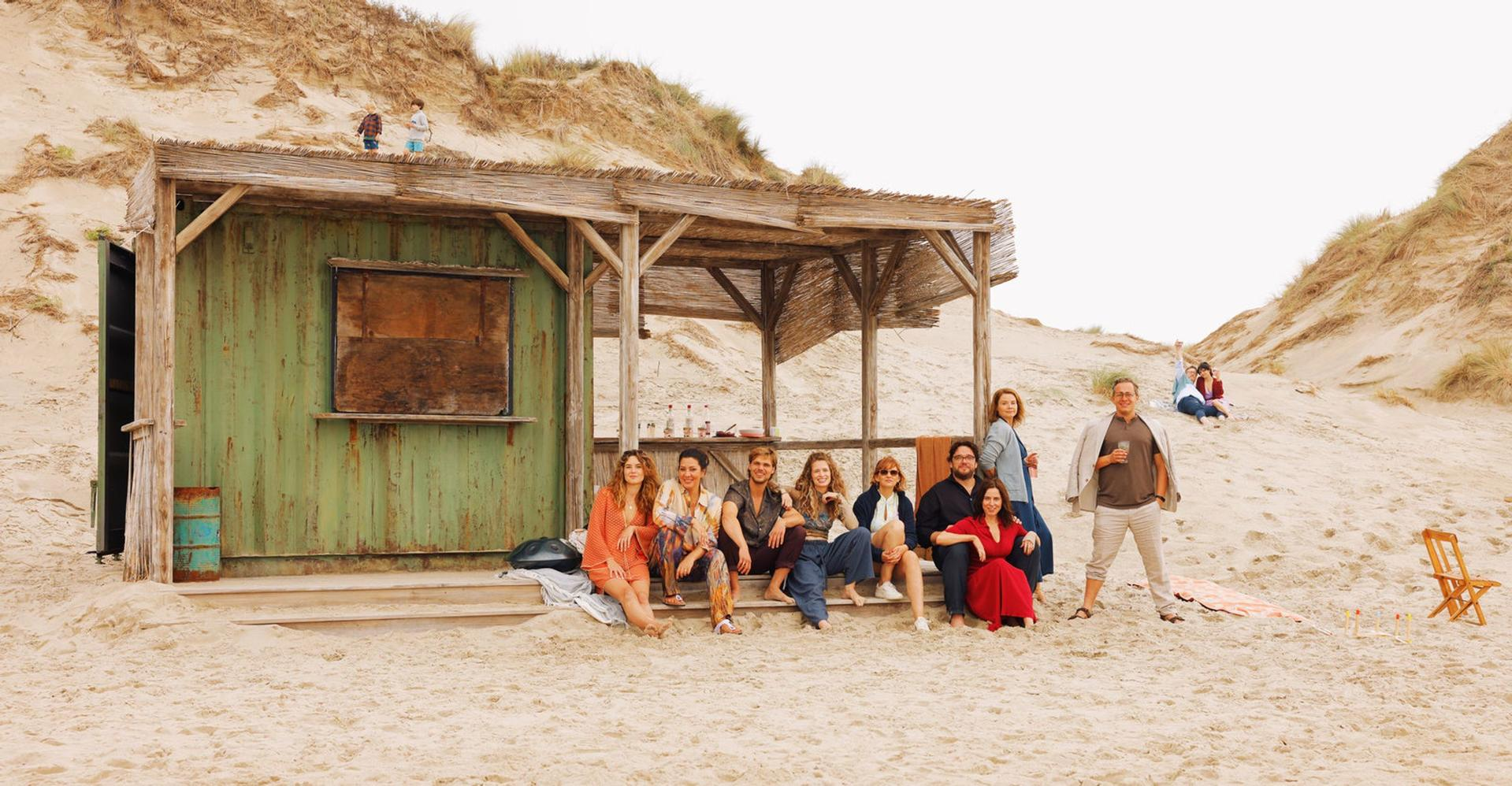 Eine Gruppe von Personen sitzt auf den Stufen einer Strandhütte. Zwei Kinder spielen auf einer Düne im Hintergrund. Ein Mann hält ein Getränk.