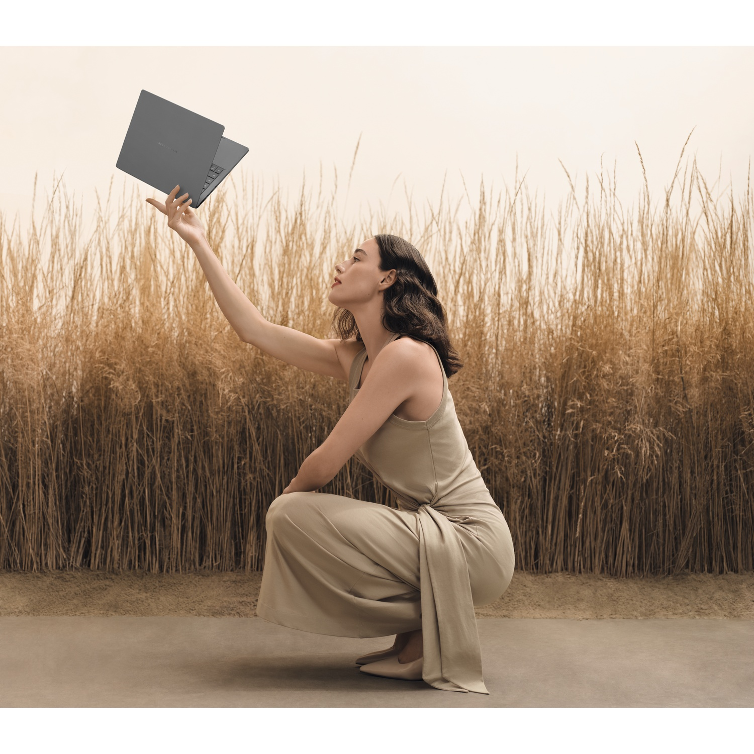 Una donna accovacciata, tiene in mano un laptop, con un campo di erba alta e secca sullo sfondo.