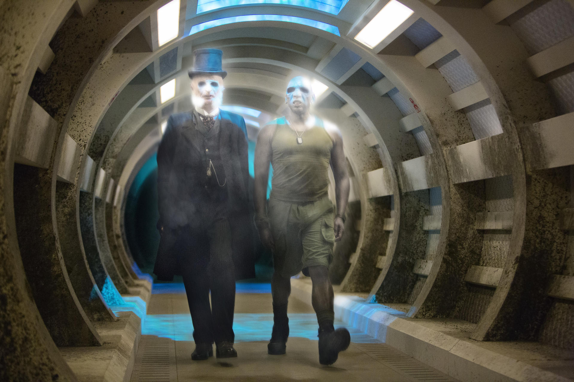Zwei gespenstische Figuren in einem Tunnel. Einer trägt einen Zylinder, der andere ein Tanktop. Der Tunnel hat ein gewölbtes Dach und blaues Licht.