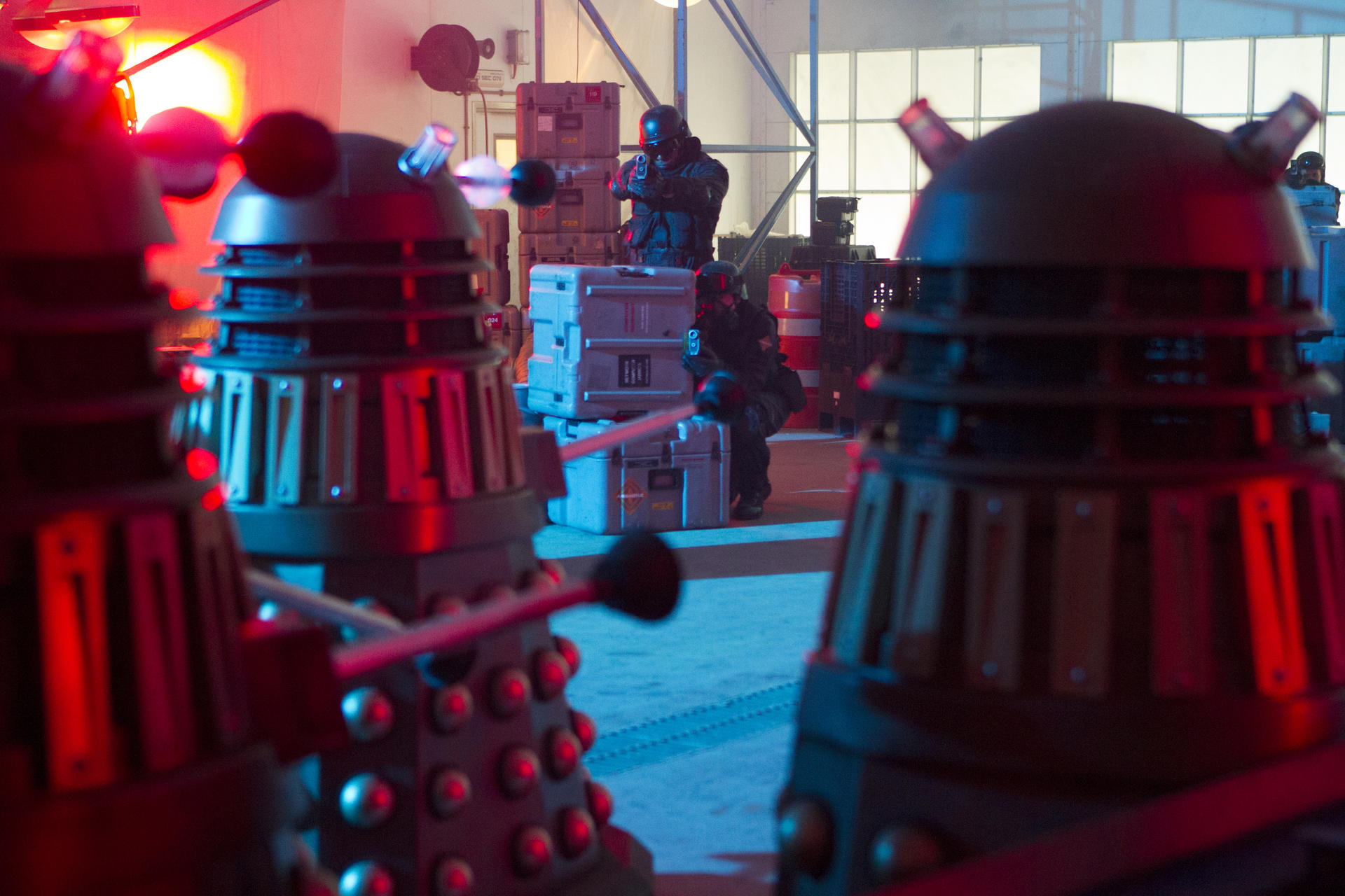 Drei Daleks in einem Lagerhaus. Zwei Soldaten zielen im Hintergrund mit Waffen. Rote und blaue Beleuchtung.