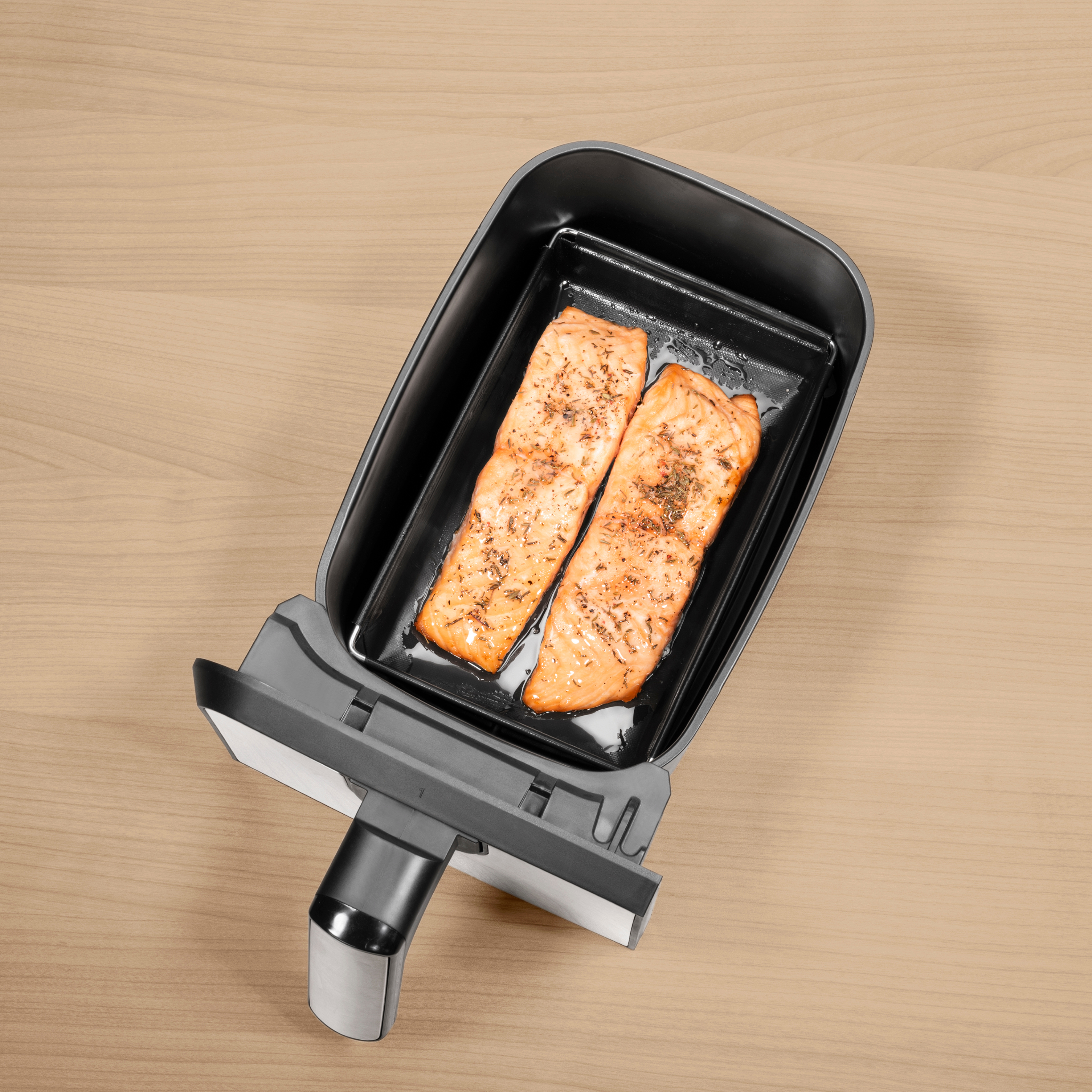 Draufsicht auf einen Airfryer-Korb mit zwei gewürzten Lachsfilets auf Holzoberfläche.