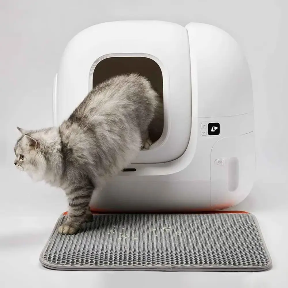 Un chat sort d'un bac à litière blanc et fermé sur un tapis gris. Sur une surface blanche.