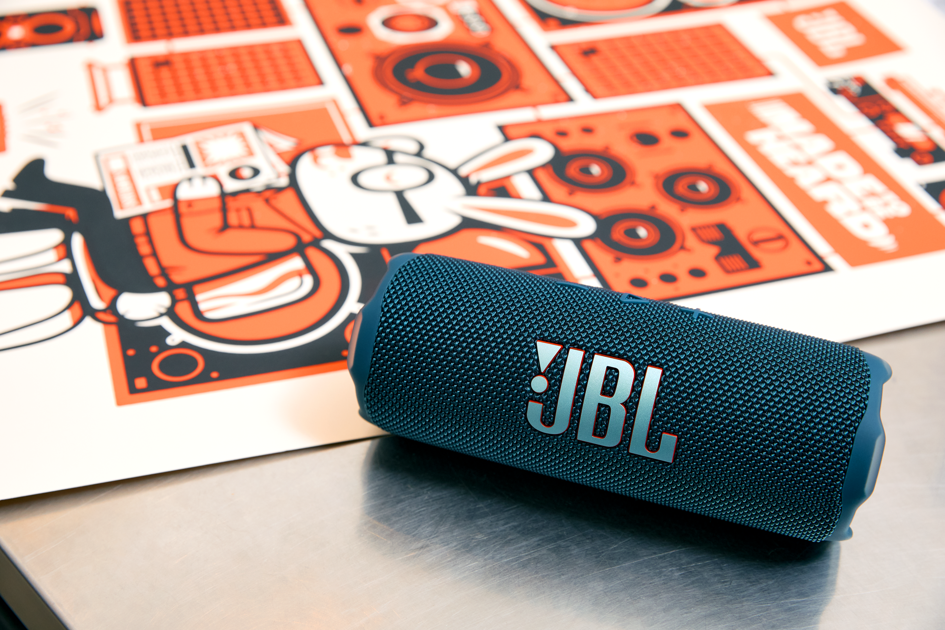 Mavi bir JBL hoparlör, kırmızı ve beyaz tasarımlı bir posterin önünde metal bir yüzeyde duruyor.