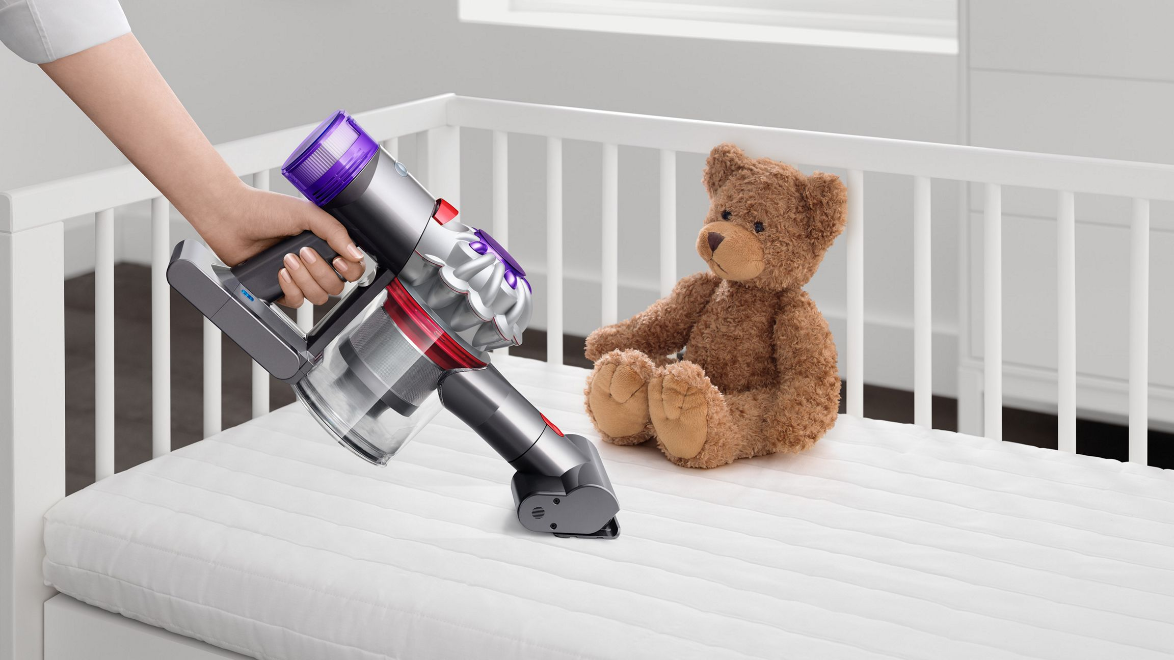 Une main tenant un aspirateur au-dessus d'un berceau blanc avec un ours en peluche.
