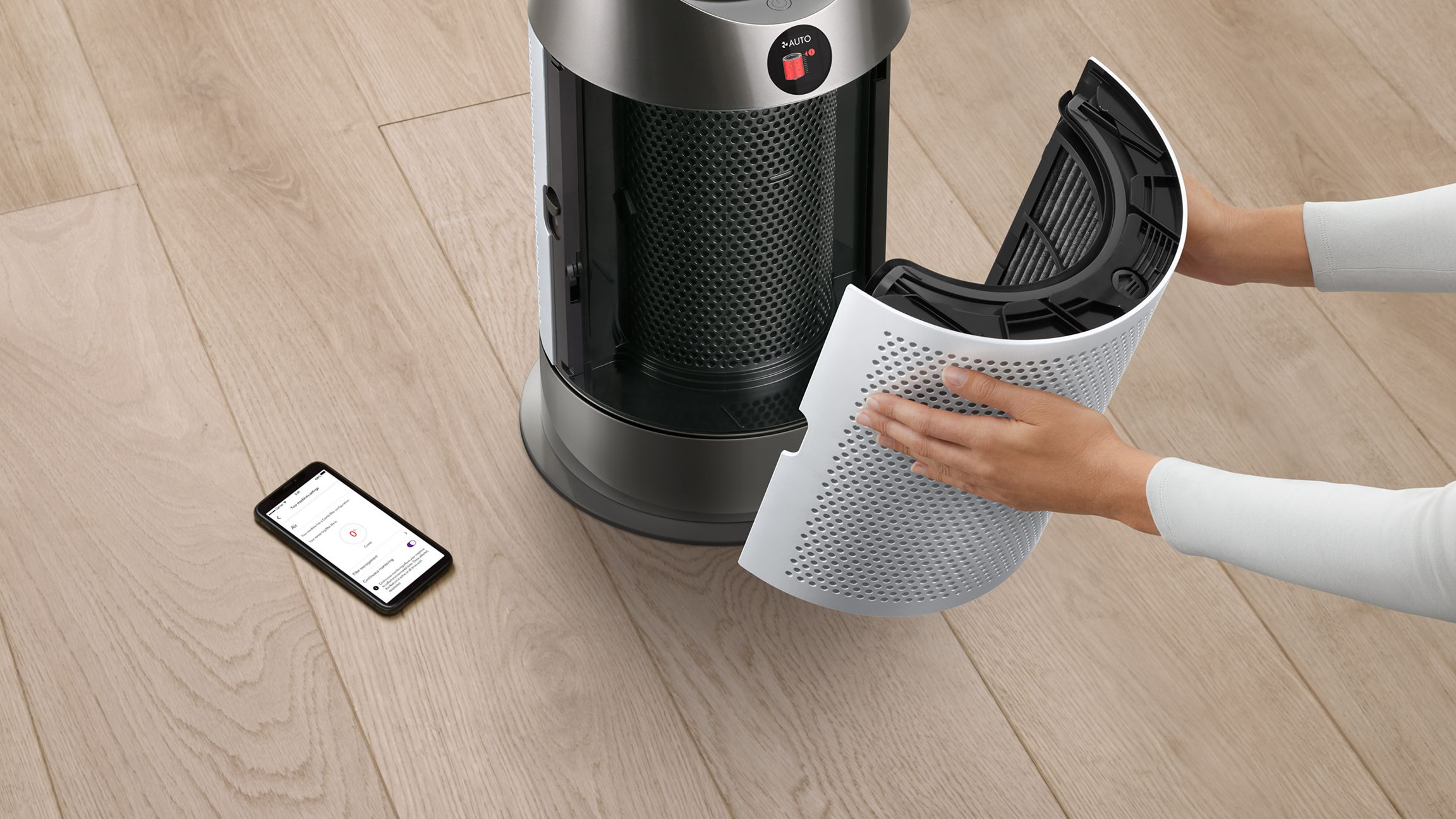 Purificateur d'air Dyson avec filtre retiré. Mains, sol en bois et smartphone visibles.