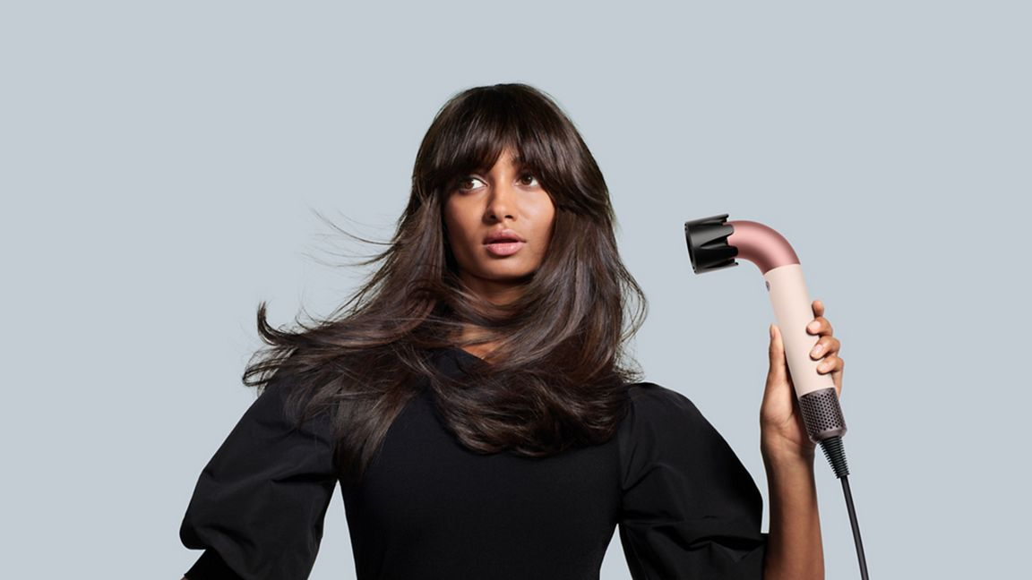 Une femme tenant un sèche-cheveux. Elle a les cheveux longs et bruns. Le fond est gris.