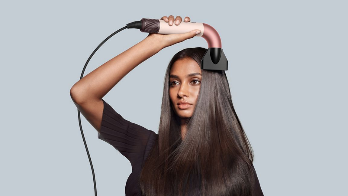 Femme aux cheveux longs utilisant un sèche-cheveux. Le sèche-cheveux est rose et noir. Elle regarde de côté.