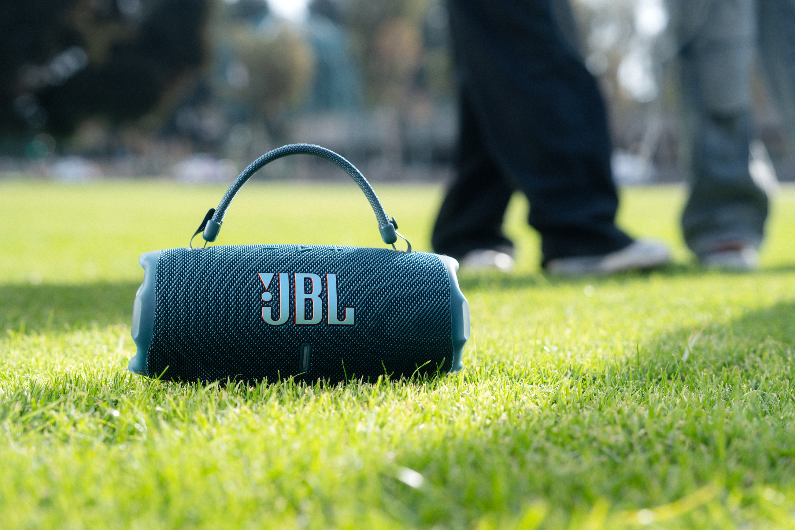 Yeşil çimenlerin üzerinde bir tutamağa sahip bir JBL hoparlör, arka planda bulanık insanlar.