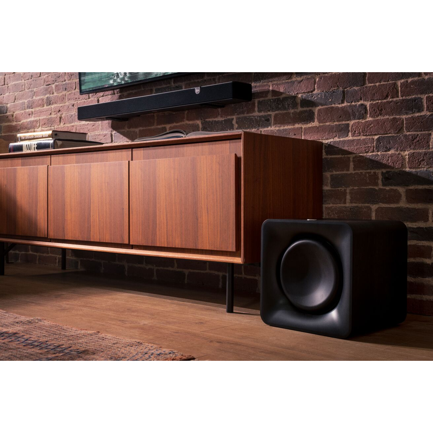 A black subwoofer next to a wooden cabinet and a brick wall. A soundbar is mounted above.