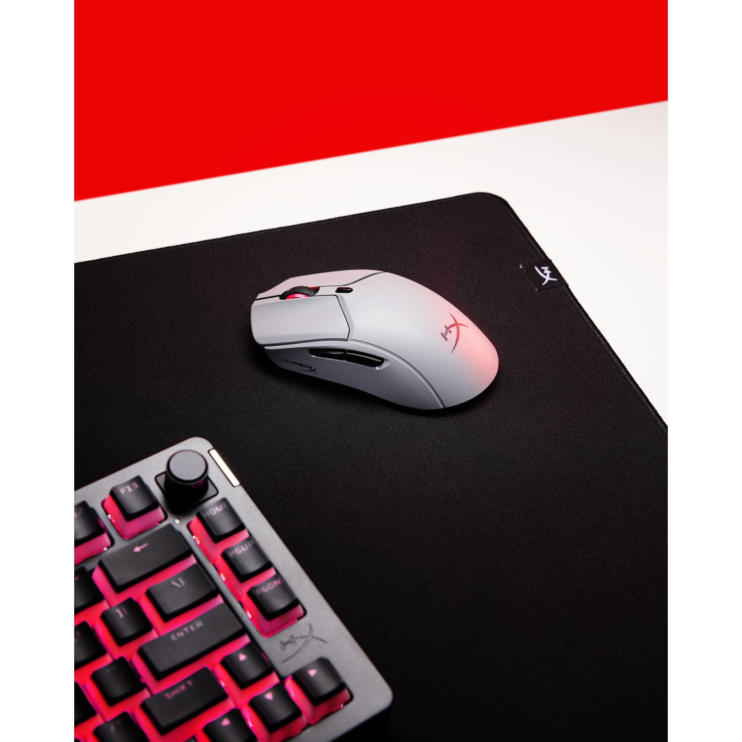 Een witte en grijze gamingmuis ligt op een zwart muismatje, naast een toetsenbord met rood verlichte toetsen.