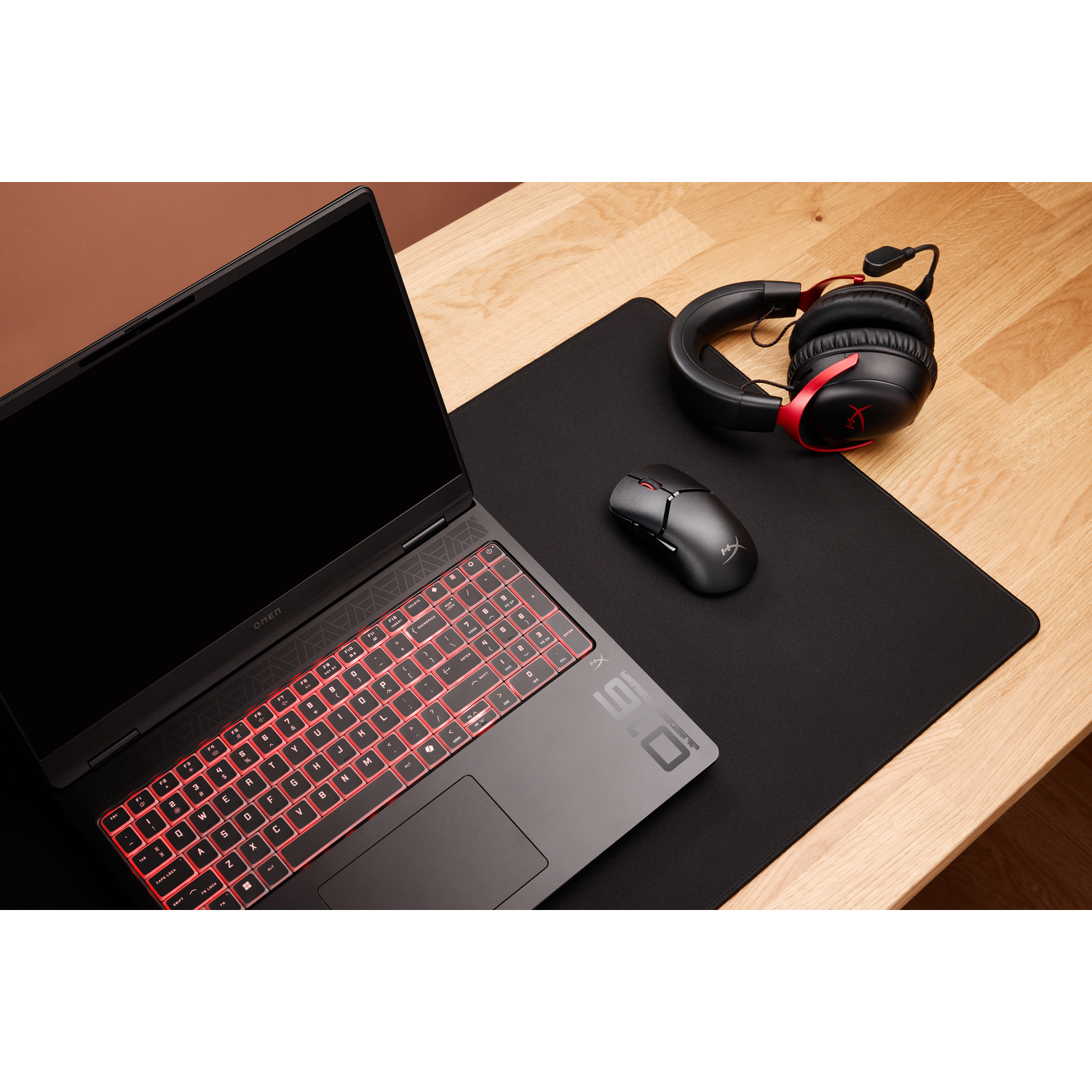 A laptop with a red-lit keyboard, a mouse, and a headset on a black mousepad and wooden table.