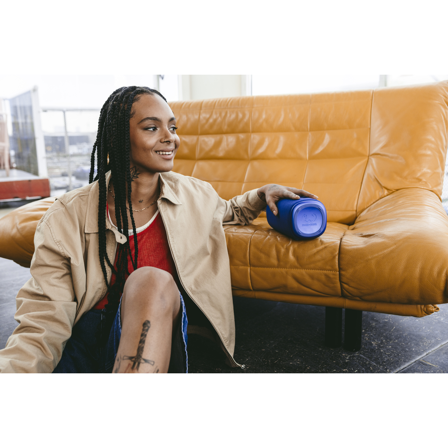 Woman holding a blue speaker, sitting on a yellow leather sofa.