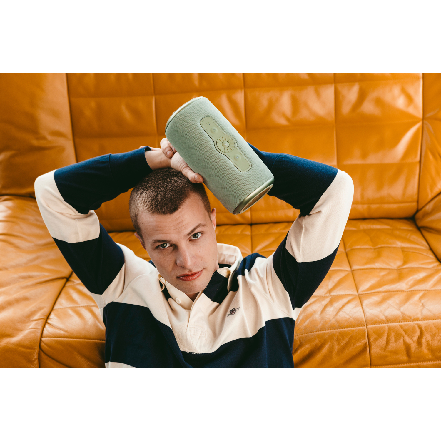 Man holding a green speaker above his head, in front of a gold couch.