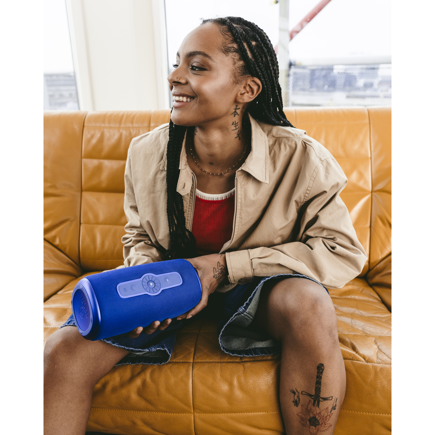 Woman with dreadlocks smiles holding a blue speaker. She is on an orange couch, wearing a jacket and shorts.