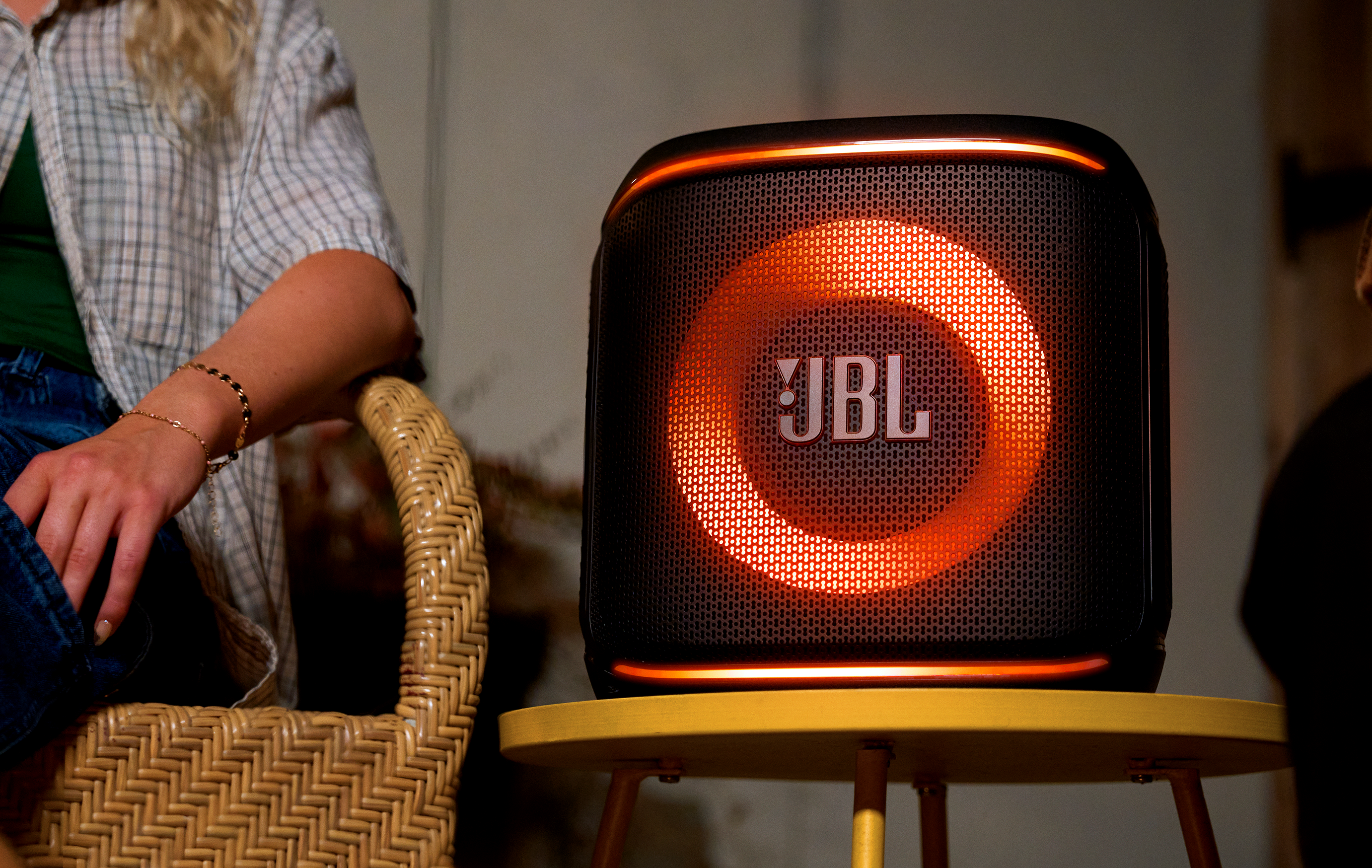 JBL hoparlör, siyah örgü ön, turuncu ışık halkası, sarı bir masanın üzerinde.