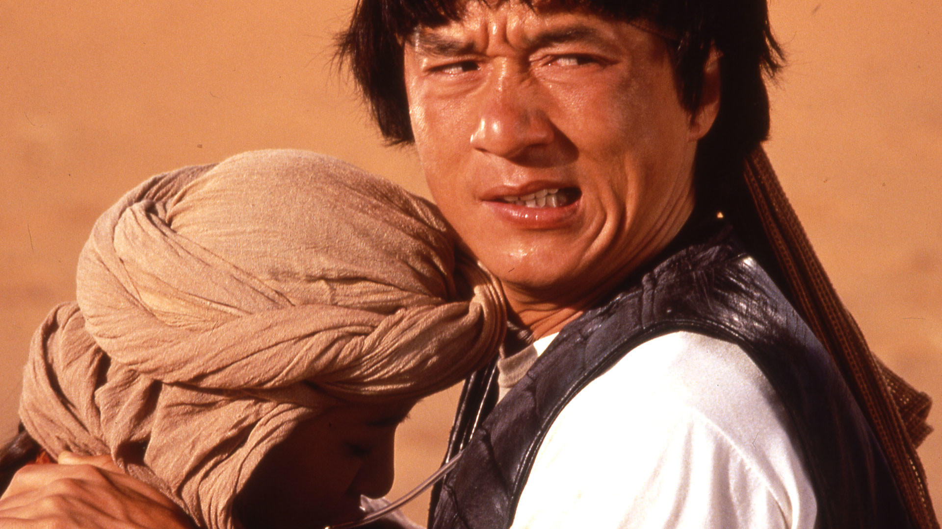 Jackie Chan umarmt ein Kind. Beide in Wüstenkulisse mit Sandsturm im Hintergrund.