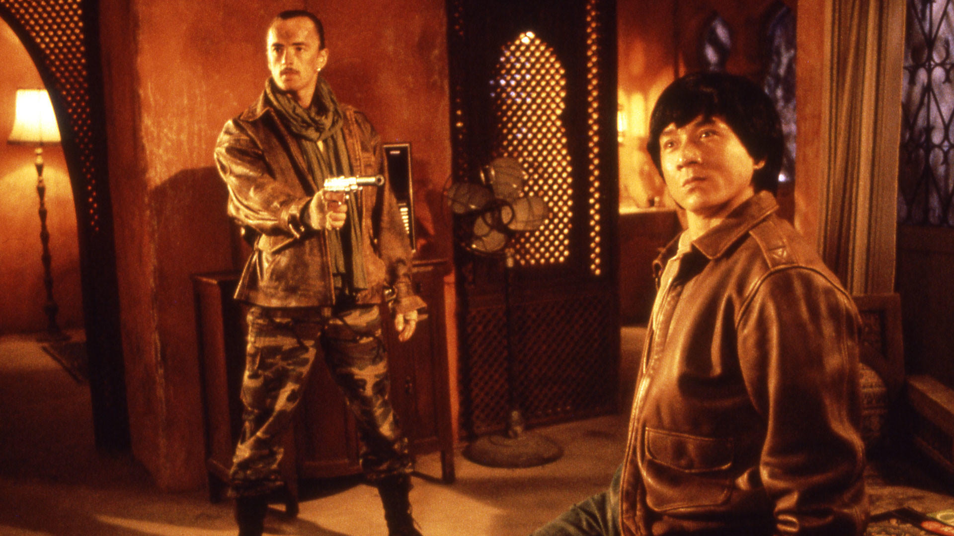 Zwei Männer in einem Raum; einer hält eine Pistole und zielt auf den anderen. Jackie Chan steht im Hintergrund und schaut zu.