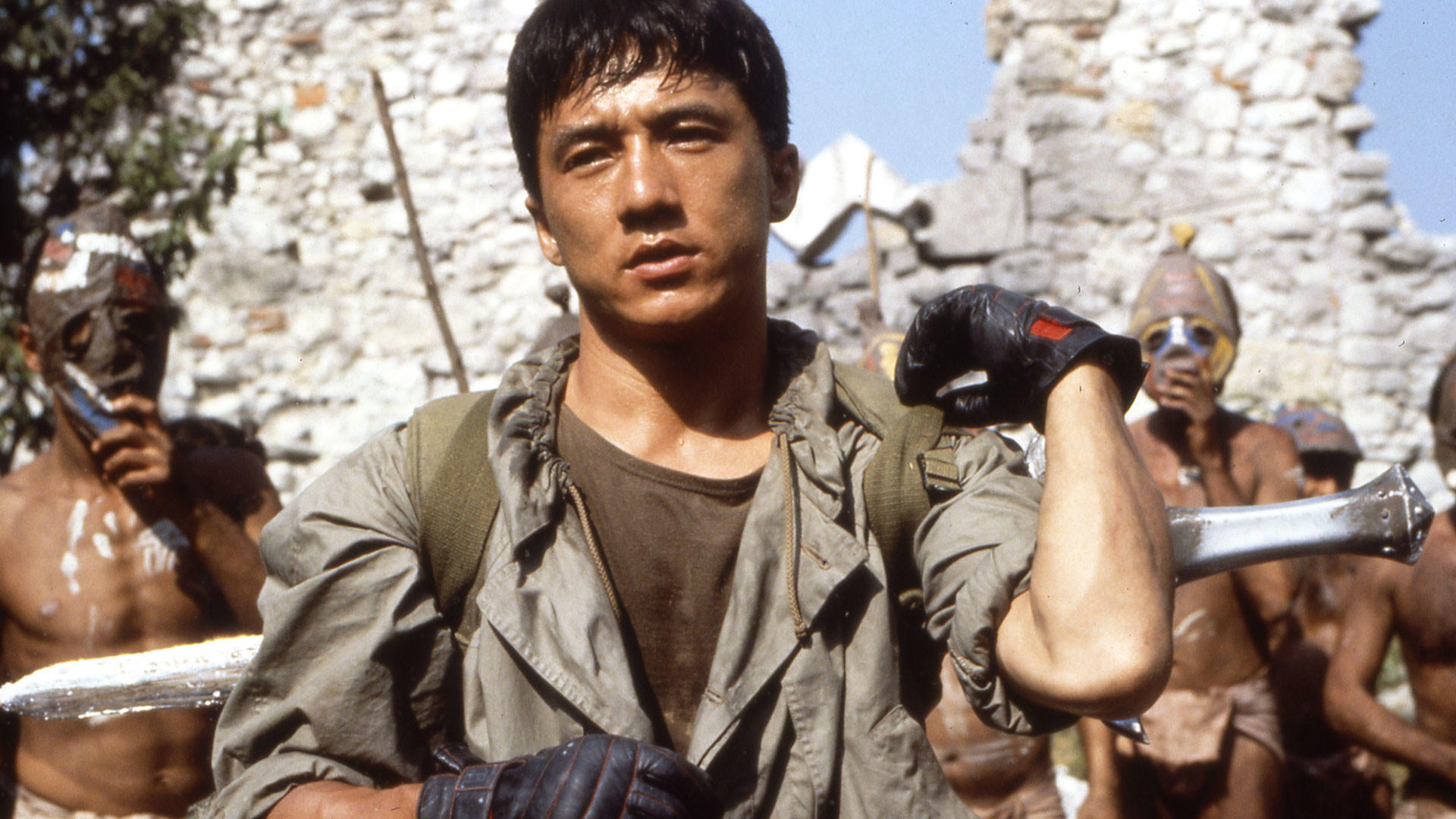 Jackie Chan hält eine Waffe und wirkt entschlossen. Andere Männer mit Stammesmasken stehen im Hintergrund, auf einer Steinmauer.