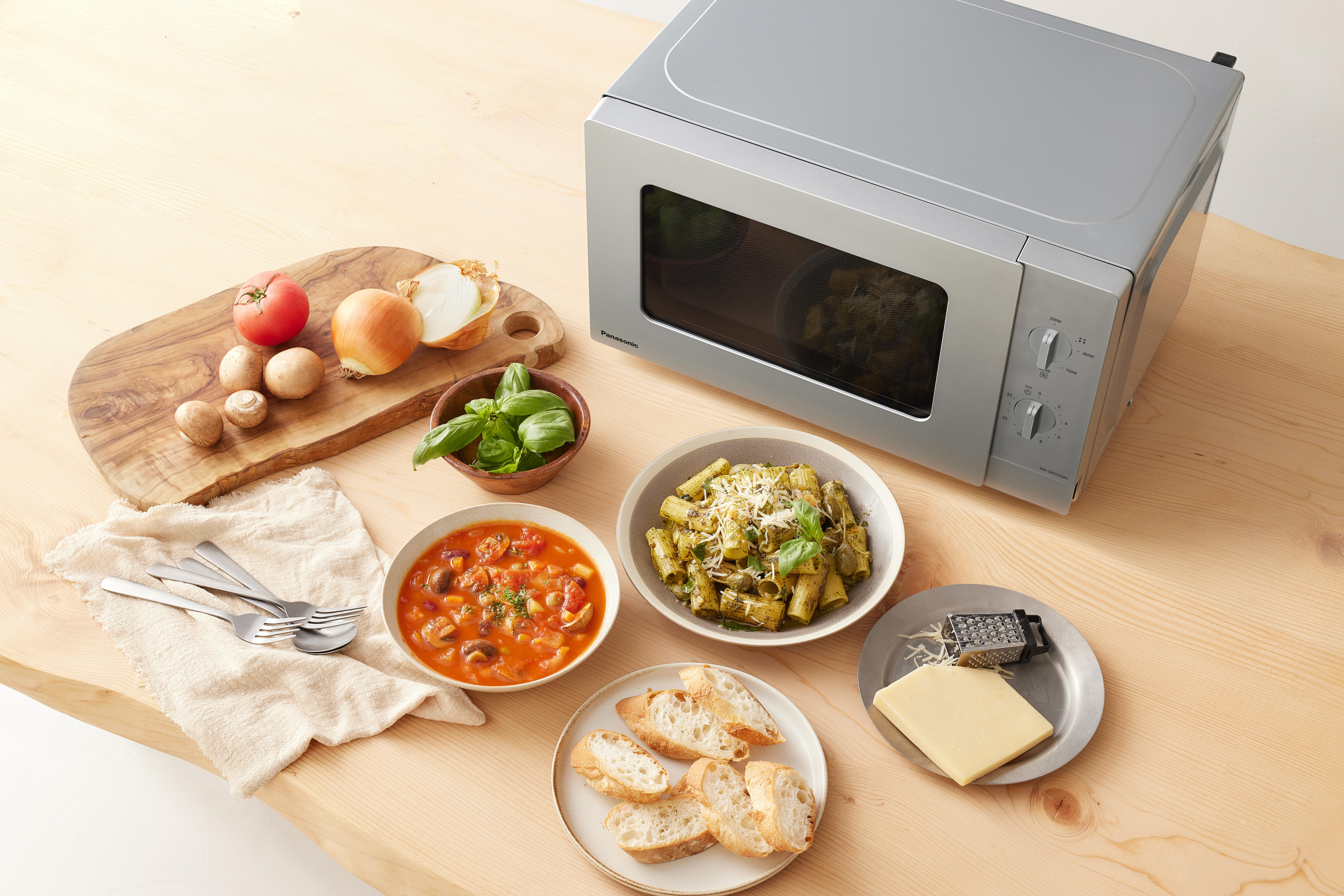 Mikrowelle mit Essen: Suppe, Pasta, Brot, Käse, Tomaten und Zwiebeln auf einem Holztisch.