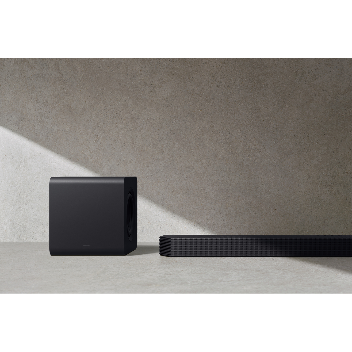 Een zwarte soundbar en subwoofer set tegen een grijze muur en vloer.