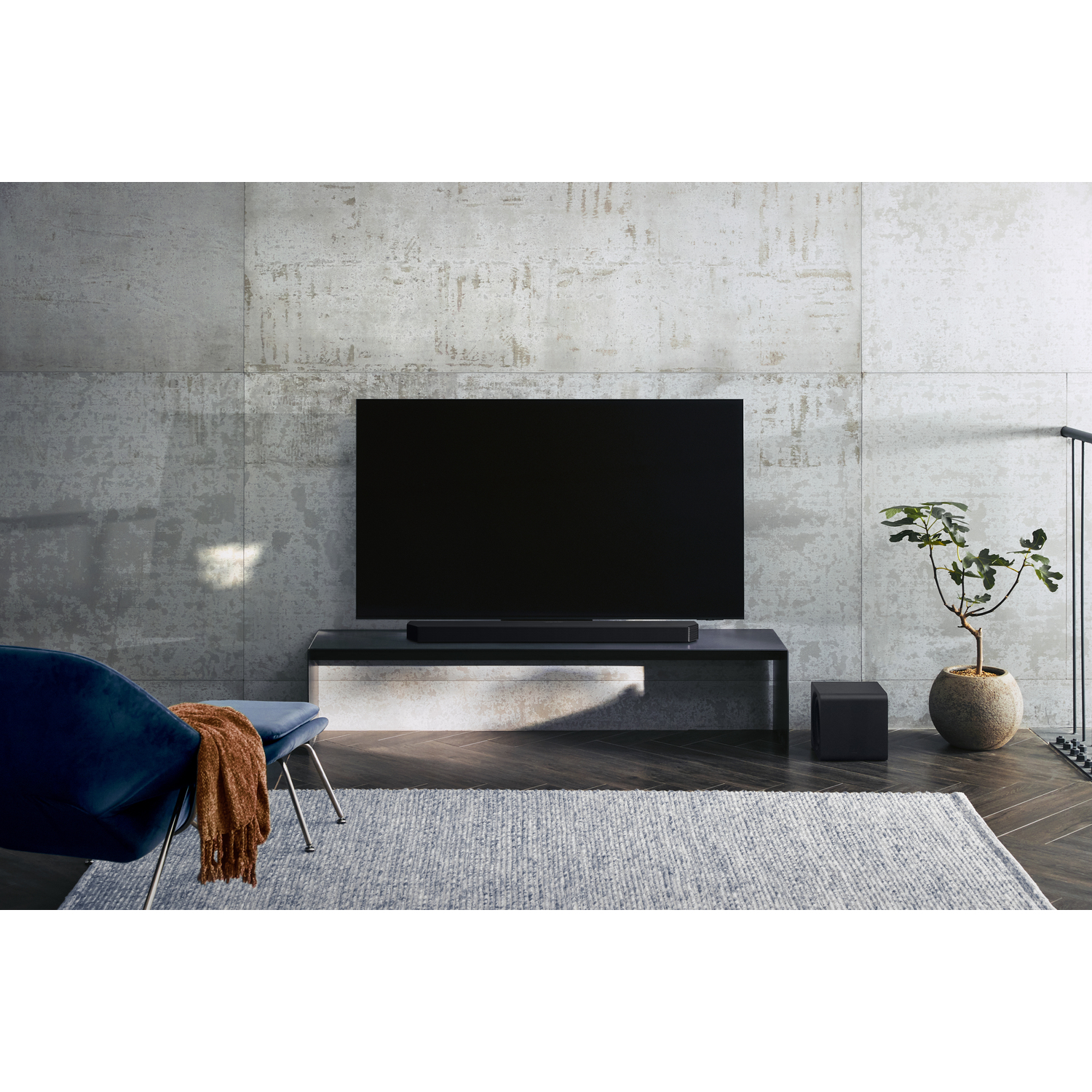 Woonkamer met tv op een standaard, soundbar, blauwe stoel, vloerkleed en plant.