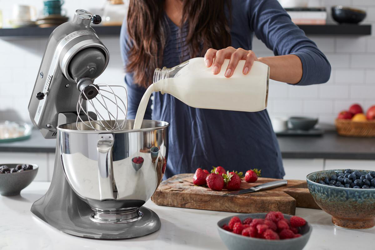 Person gießt Milch in eine graue Küchenmaschinen-Schüssel, Erdbeeren und Blaubeeren daneben.