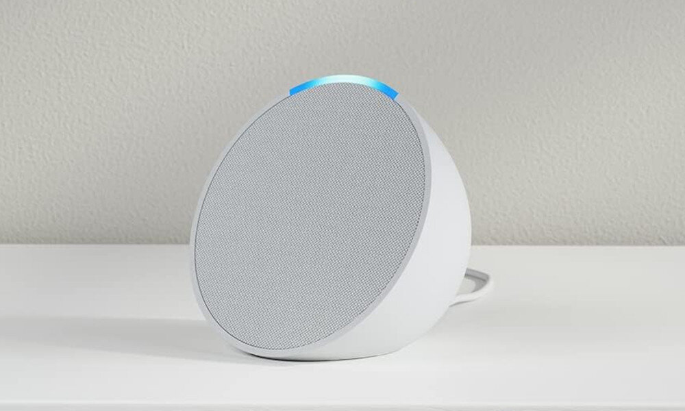 Biały głośnik inteligentny z szarą siatką głośnika, niebieskim światłem na górze, na białej powierzchni na białej ścianie.