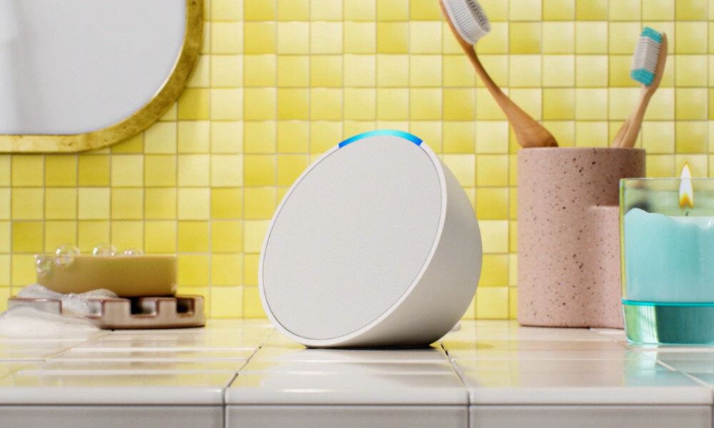 Biały głośnik smart, mydło, szczoteczki do zębów, świeca w łazience z żółtą kafelkową ścianą i lustrem.