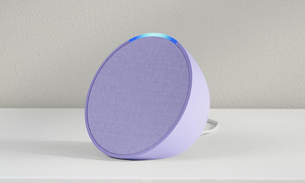 Fioletowy głośnik na białej powierzchni. Góra ma niebieskie światło. Białe tło.