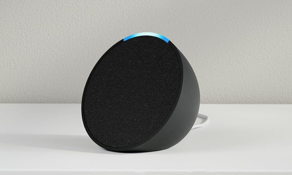 Czarny głośnik z siatkowym przodem, niebieskim światłem na górze, na białym stole.